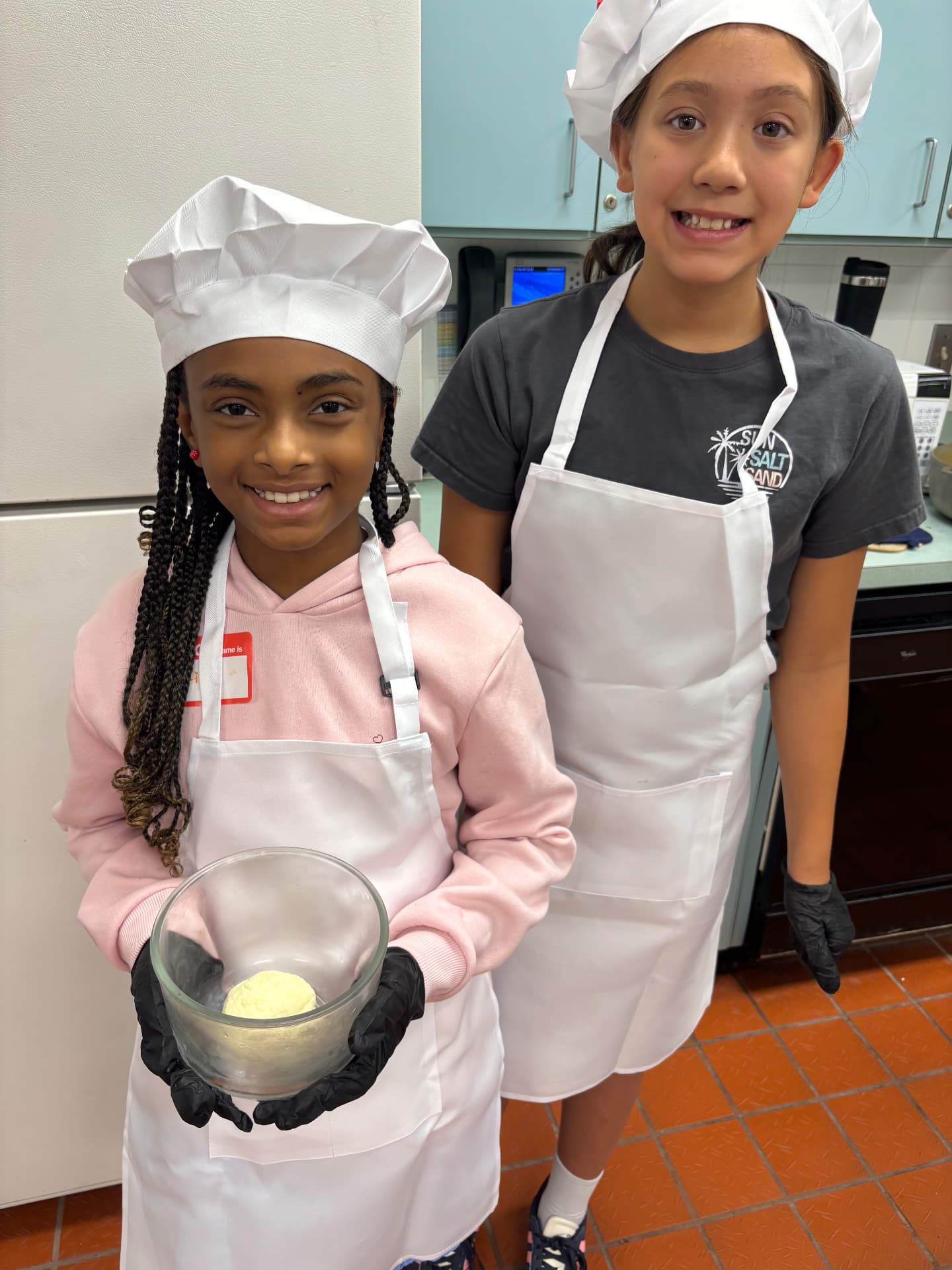 Two young chefs in hats and aprons proudly showing dough they prepared together